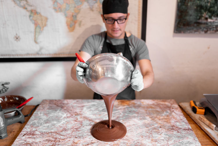Hispanic man in uniform and glasses pouring liquid chocolate from bowl on marble board during work in confectionery