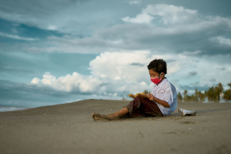 Boy wearing mask writing in paper while sitting at beach against sky