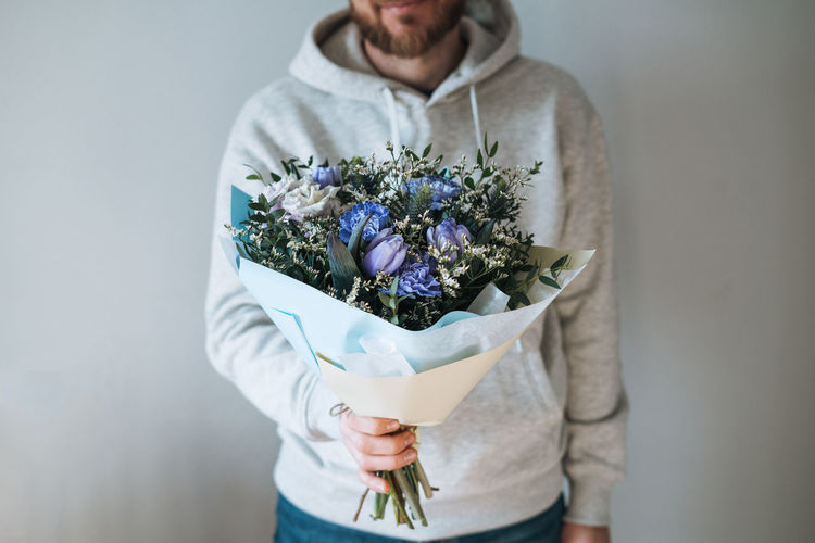 Young bearded man in grey hoodie with bouqet of flowers in hands on the grey background