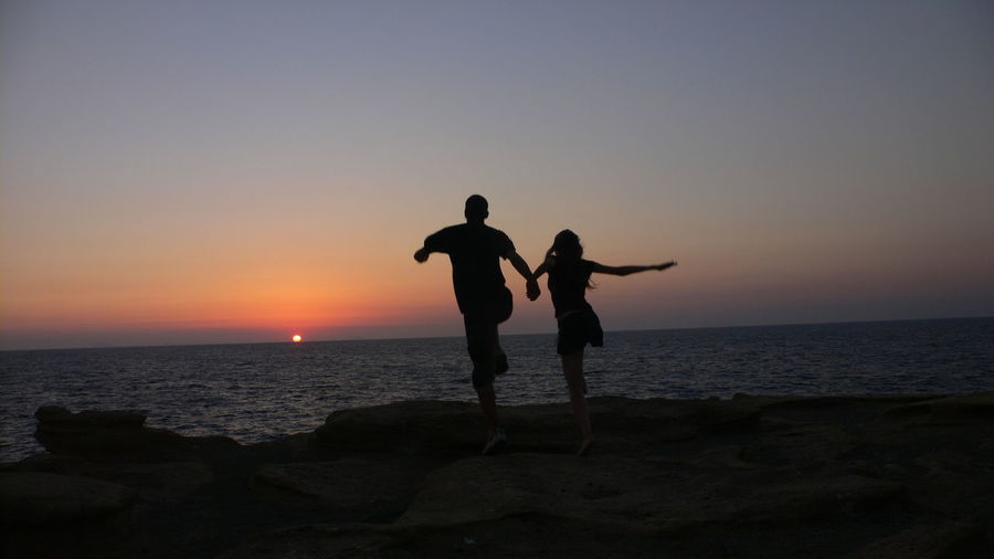 Ear view of couple jumping at rocky seashore against sky during sunset