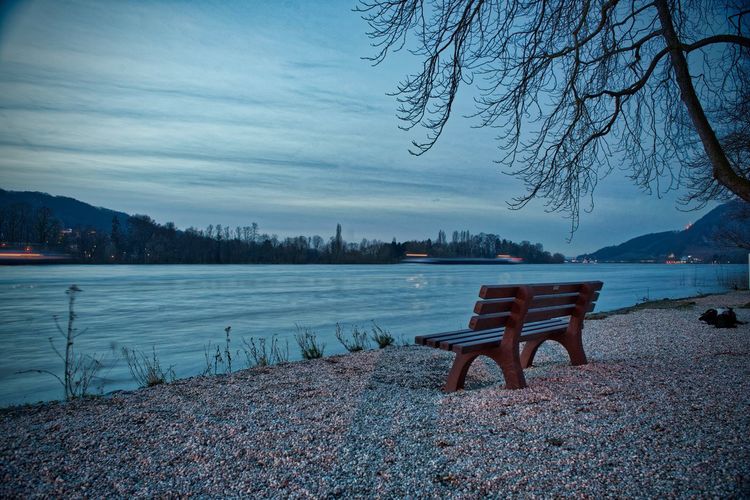 Empty bench by lake against sky during winter