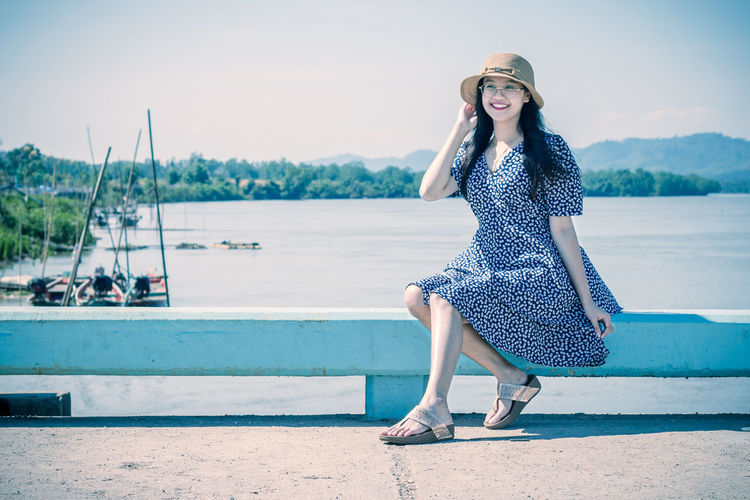 Full length of smiling young woman sitting on railing against lake