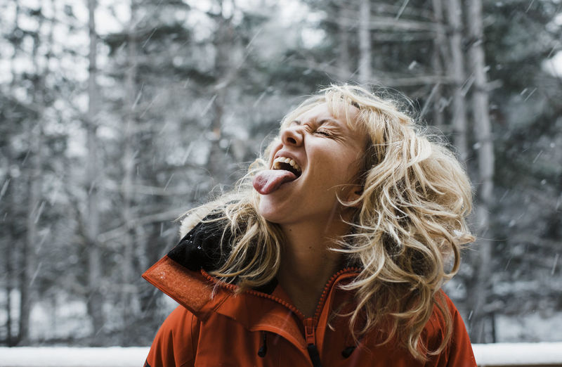 Woman with eyes closed sticking out tongue while standing in forest during snowfall