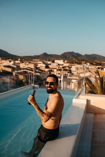 Side view of cheerful young shirtless ethnic male traveler with dark hair and beard in shorts and sunglasses smiling while standing in swimming pool and drinking beer on rooftop