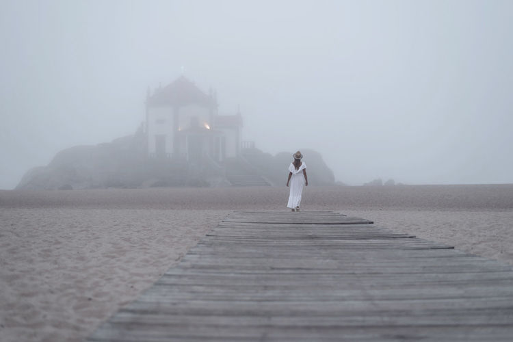 Back view of unrecognizable feminine tourist standing admiring aged chapel of senhor da pedra and ocean in fog from pier in porto portugal