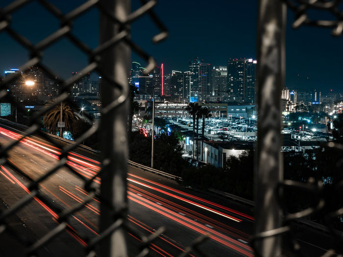 High angle view of light trails on road in city seen through broken chainlink fence at night
