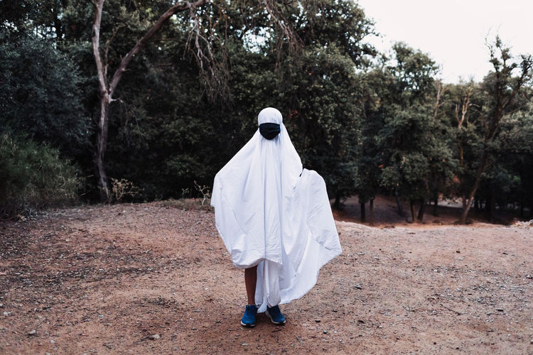 Faceless creepy child in ghost costume standing in park during halloween on overcast day