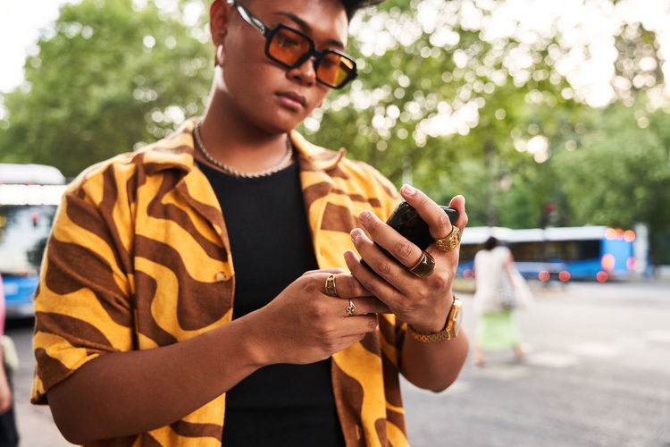 Modern asian male in trendy orange shirt and sunglasses checking smartphone while standing on street