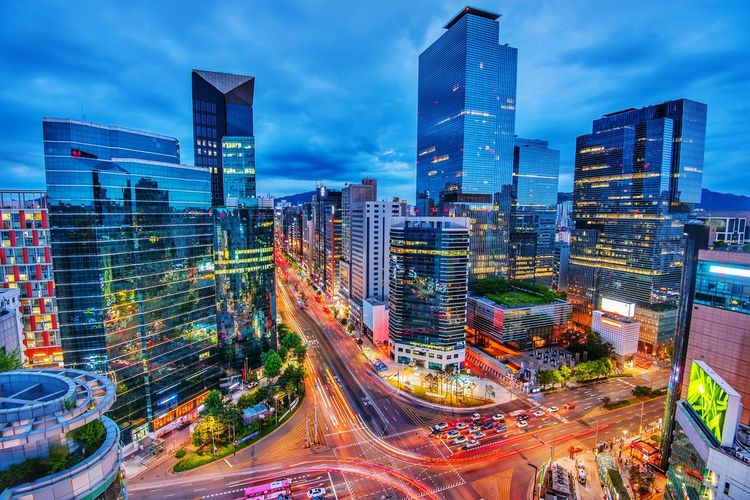 Illuminated city street by buildings against sky at dusk