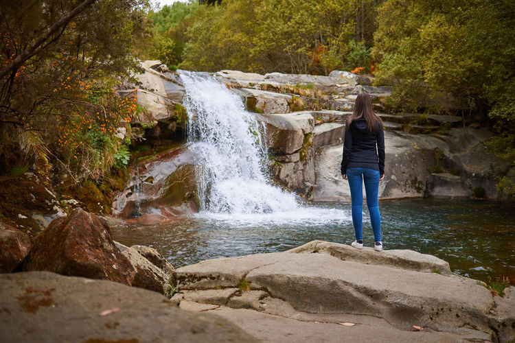 Full length rear view of woman standing on rock against waterfall