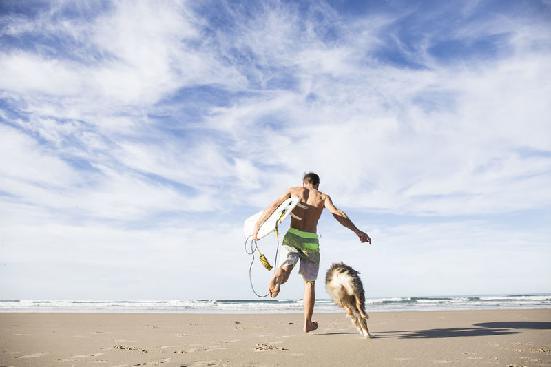 Man carrying surfboard running with dog on the beach