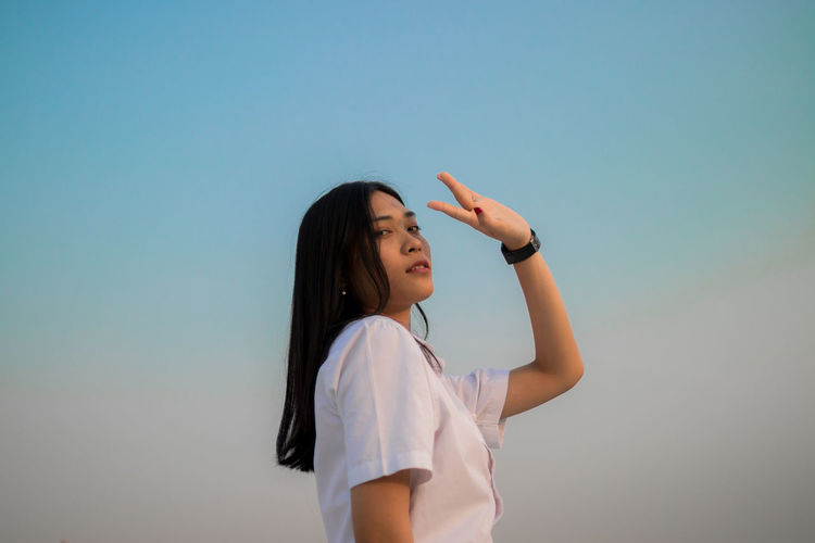 Low angle view portrait of young woman shielding eyes standing against sky during sunset
