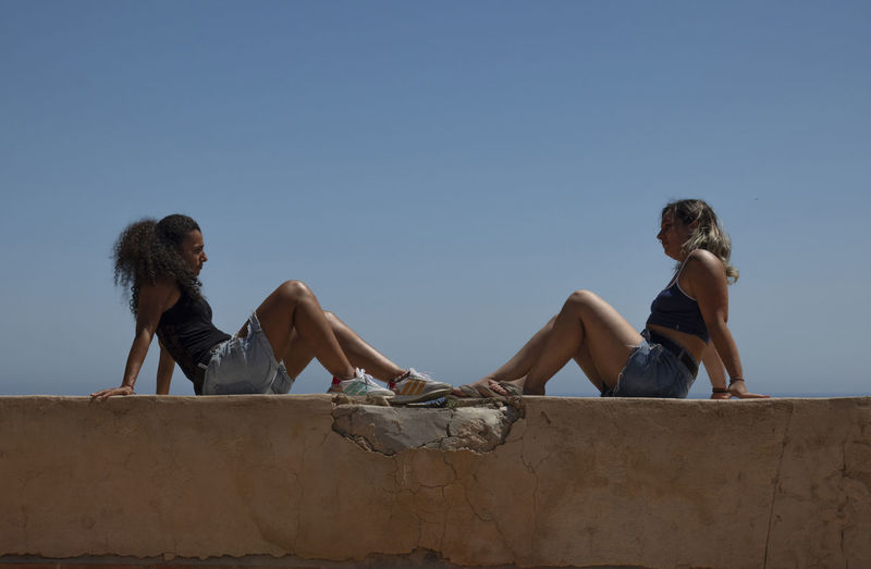 Low angle view of 2 girls sitting on wall beside the sea against clear sky