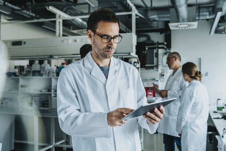 Scientist wearing eyeglasses using digital tablet while standing with coworker in background at laboratory