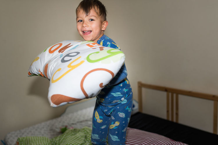 Portrait of happy boy holding pillow while standing on bed at home