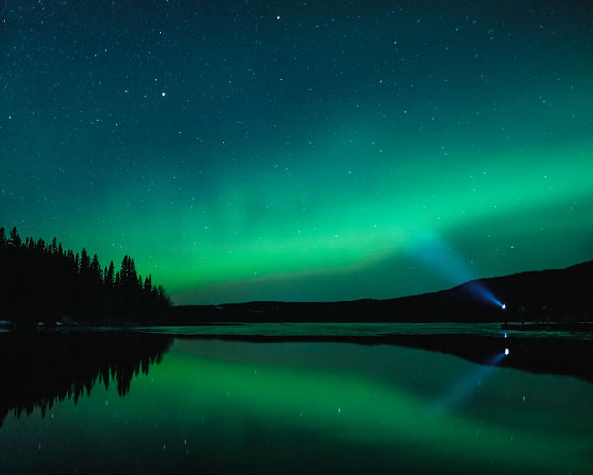 Man standing at clear reflecting lake with head lamp shining into aurora filled night skies