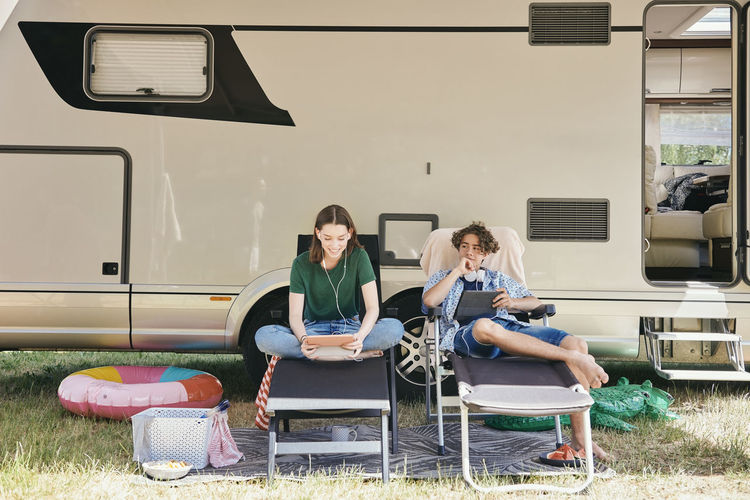 Brother and sister using digital tablet while sitting on folding chairs against motor home