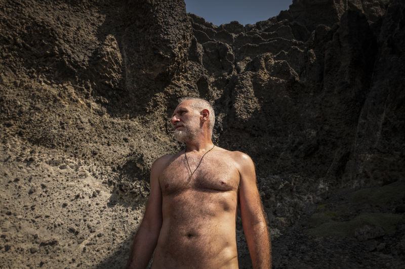 High angle view of portrait of shirtless adult man on beach against rock
