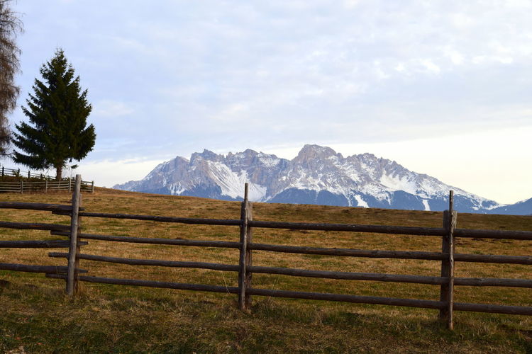 Fence on field by snowcapped mountain against cloudy sky