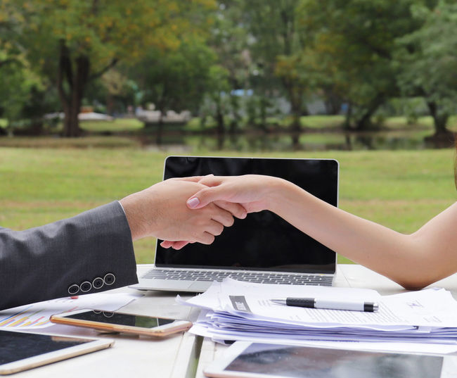 Cropped image of colleagues handshaking by laptops and document in park