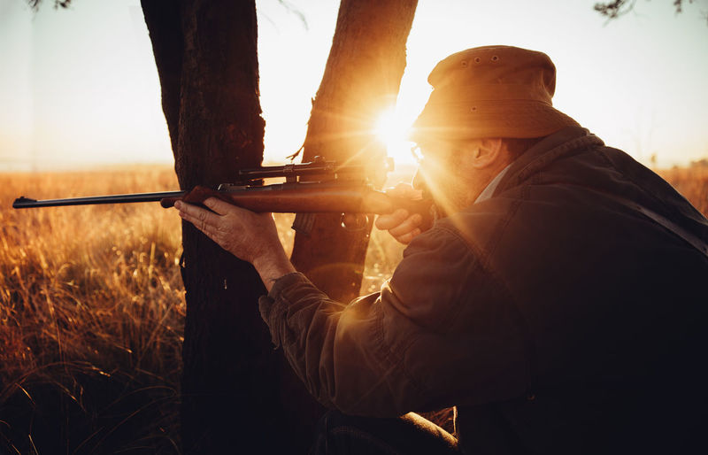 Man aiming rifle by tree trunk on field