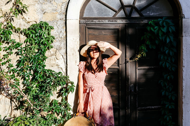 Beautiful young woman in pink dress standing in front of picturesque old house, door, light, shadow.