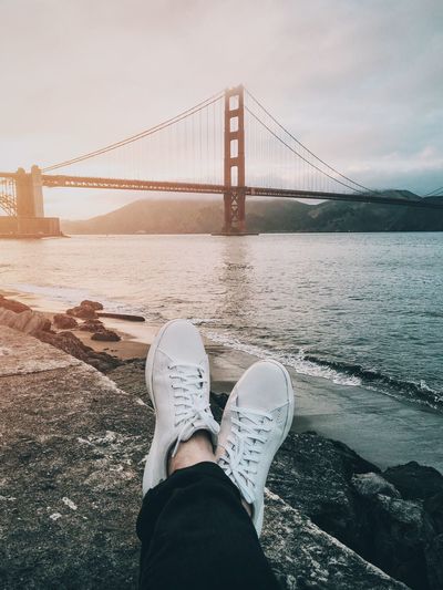 Low section of person wearing shoes while sitting by river against bridge 