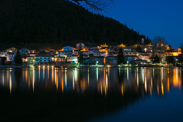 Illuminated buildings by lake against sky at night