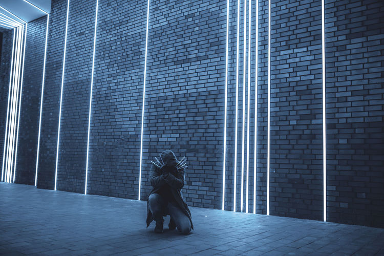 Full length of woman holding illuminated sticks while kneeling on footpath against brick wall
