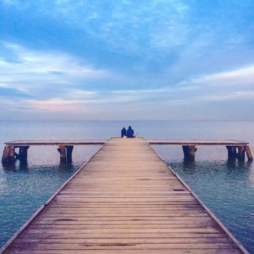 People sitting on edge of pier by sea against cloudy sky