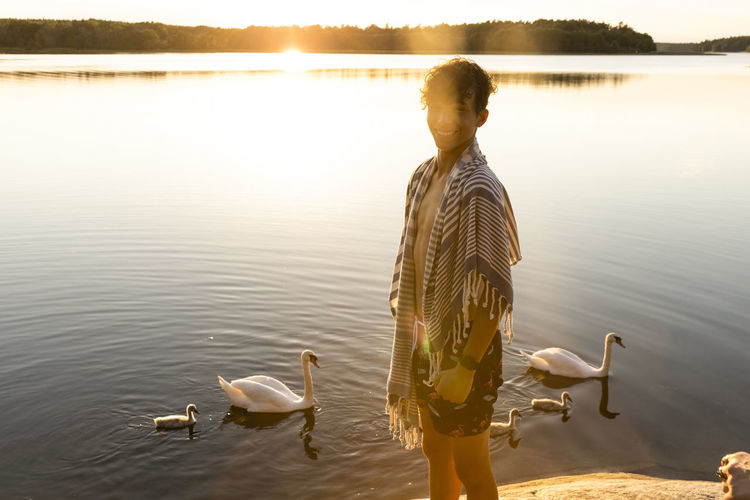Portrait of happy boy standing near lake with swans swimming in background