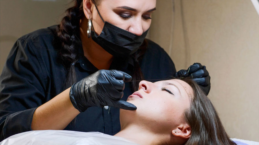 Close-up of beautician wearing mask threading woman in salon