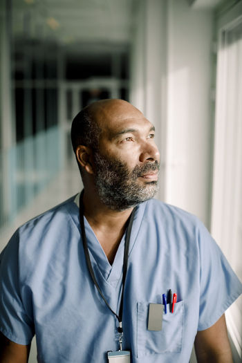 Contemplating male nurse looking through window while standing in hospital corridor