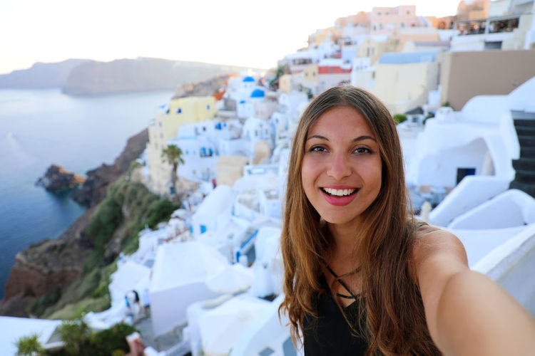 Portrait of smiling young woman standing against buildings at oia in santorini