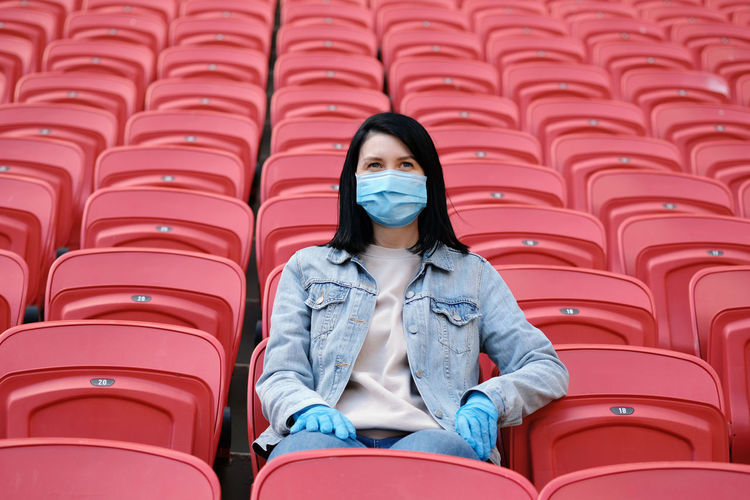 A female fan in a medical mask and rubber gloves sits alone in an empty stadium with red seats.