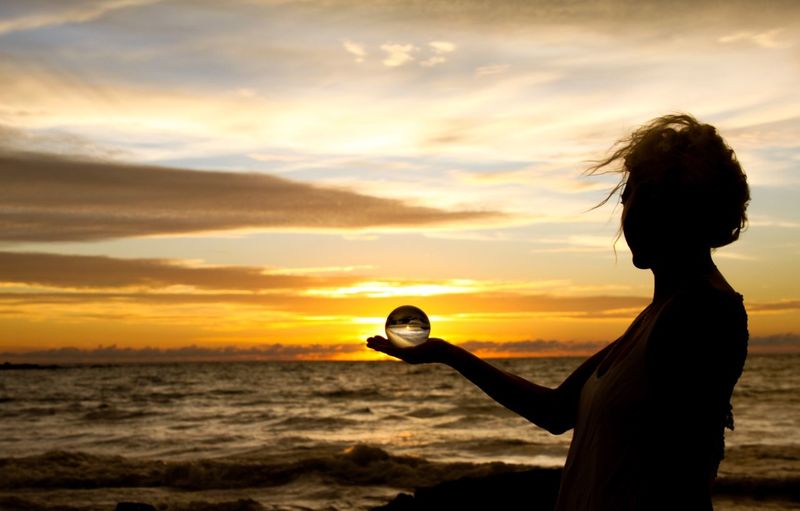 Woman holding crystal ball while standing at beach against sky during sunset