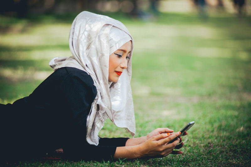 Side view of woman wearing hijab while using mobile phone on grassy field at park
