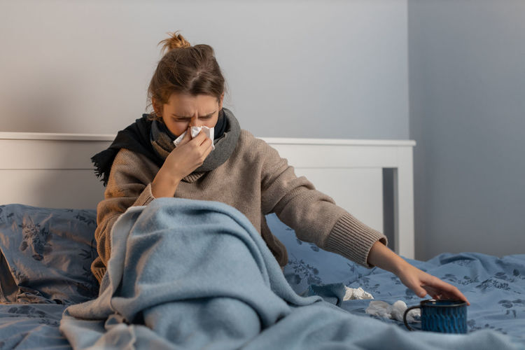 Cute woman blowing nose into a paper napkin and pulls hand to cup with warm beverage. 