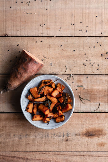 Top view of half of raw sweet potato and pieces of baked sweet potato in bowl on rustic wooden table