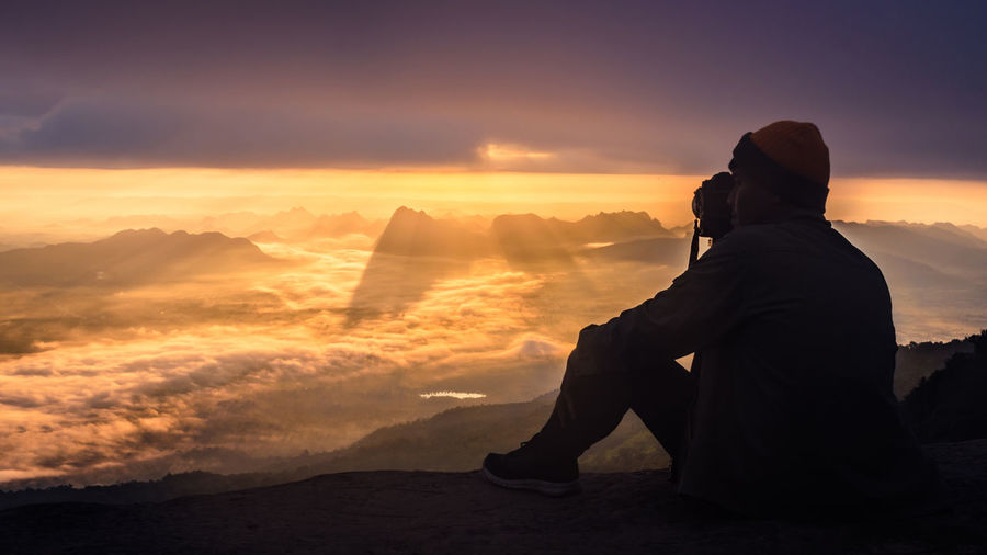 Rear view of silhouette man sitting on mountain against sky during sunset