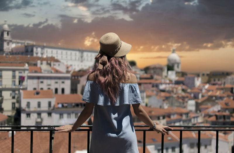 Rear view of young woman looking at cityscape while standing by railing in balcony against sky during sunset