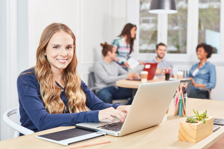 Portrait of cheerful businesswoman using laptop with colleagues in background at office