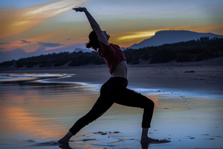 Full length of young woman doing yoga at beach against sky during sunset