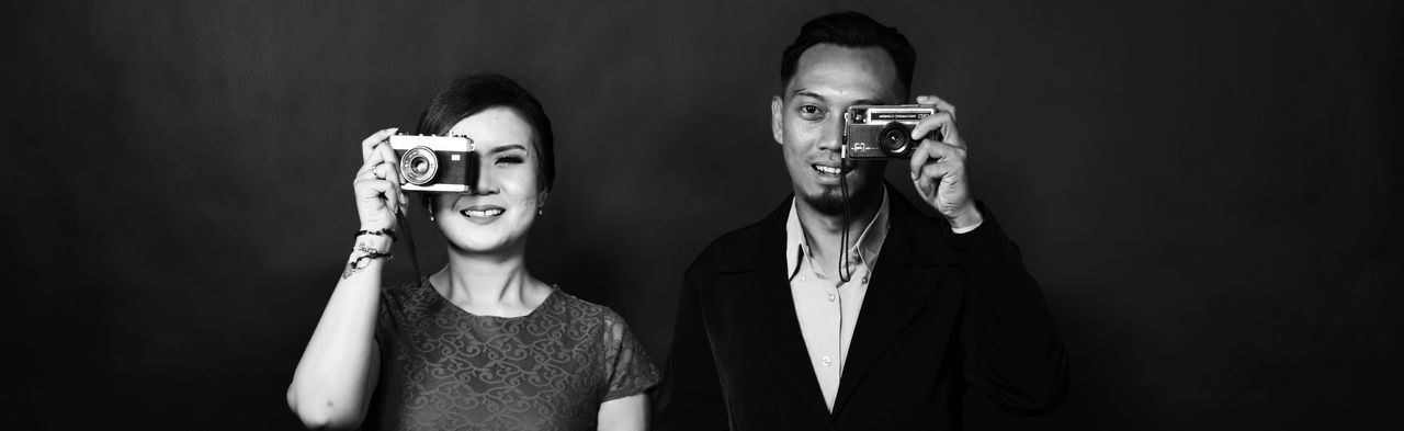 Portrait of smiling man and woman holding old cameras against black background
