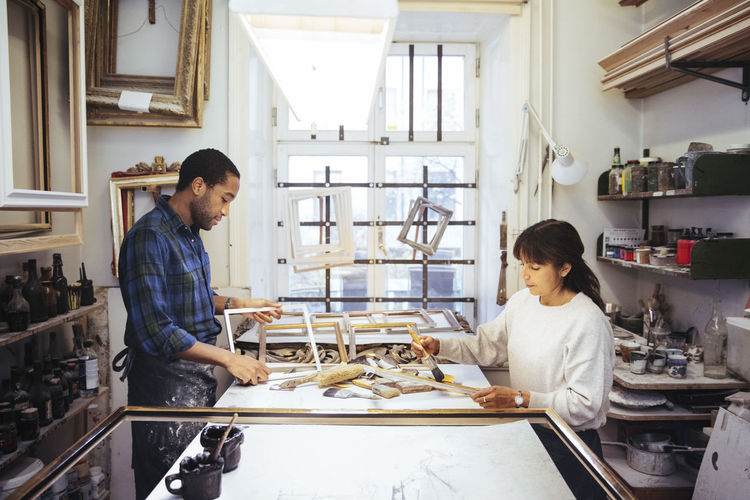 Male craftsperson making frame with female coworker at workbench in store