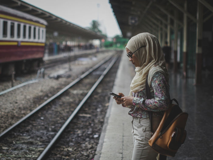 Side view of woman using phone while standing at railroad station