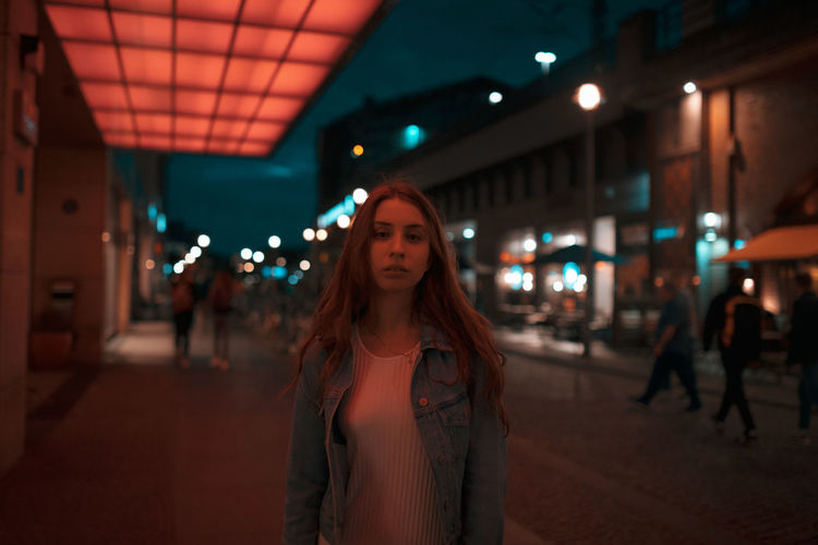 Portrait of woman standing on footpath under illuminated lights