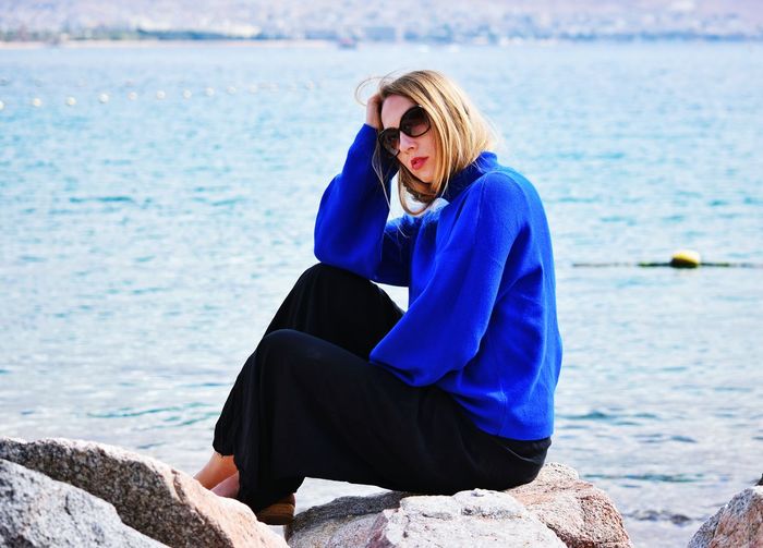 Woman wearing sunglasses sitting on rock by sea against sky