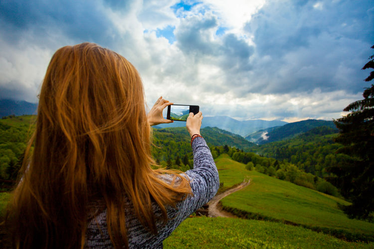 Rear view of woman photographing while standing on landscape against cloudy sky