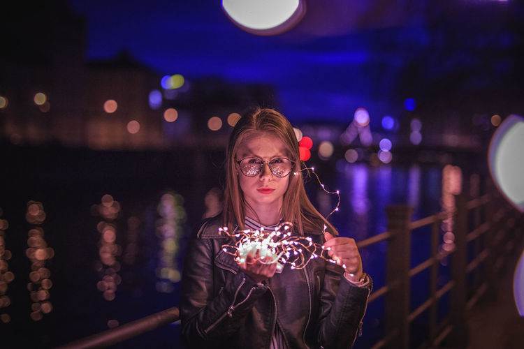 Portrait of young woman holding illuminated string lights by lake at night